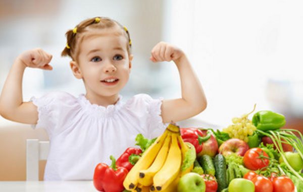 The girl shows how strong she is. There are fruits and vegetables on the table 