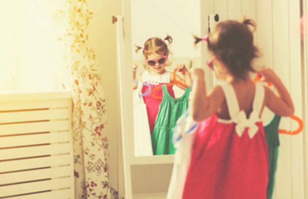 Маленькая девочка в солнцезащитных очках стоит перед зеркалом, держа в руках вешалки с платьями